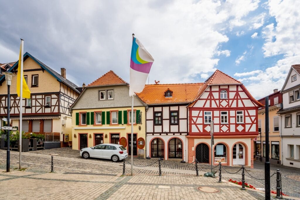 Ein an einem Hang angelegter, gepflasterter Platz. Dahinter an einer ebenfalls gepflasterten Straße vier Fachwerkhäuser in unterschiedlichen Farben. Vor einem der Häuser steht ein weißer PKW.