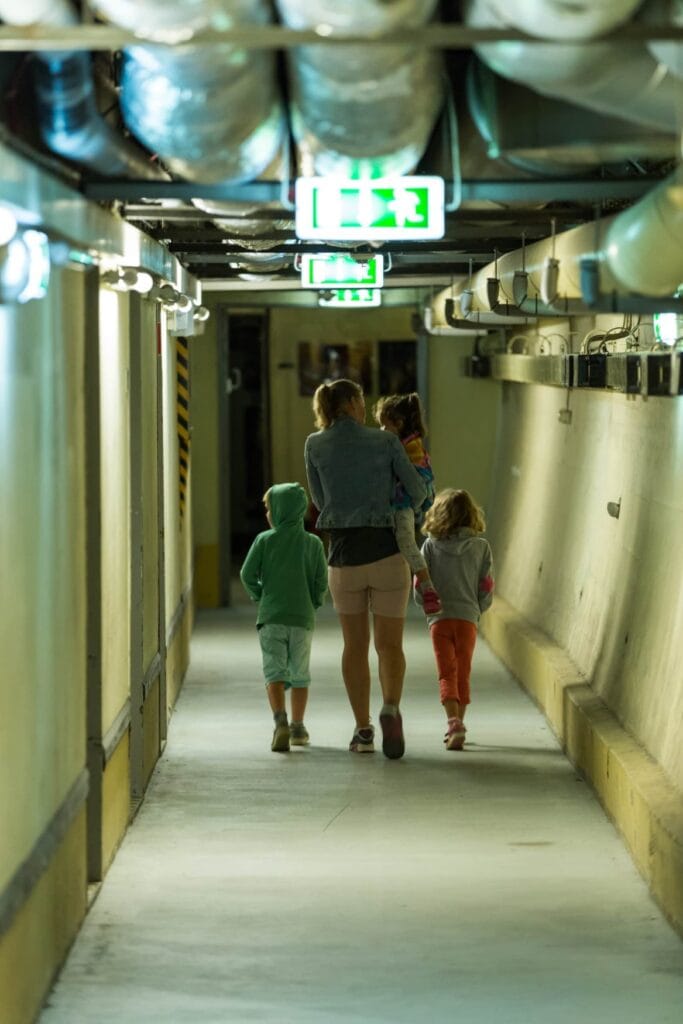 Eine Frau mit zwei Kindern und einem dritten Kind im Arm läuft geradewegs von der Kamera weg durch einen unterirdischen, beleuchteten Gang eines Bunkers. An der Decke verlaufen mehrere Rohre und Kabel, in regelmäßigen Abständen sind Notausgangsschilder angebracht.