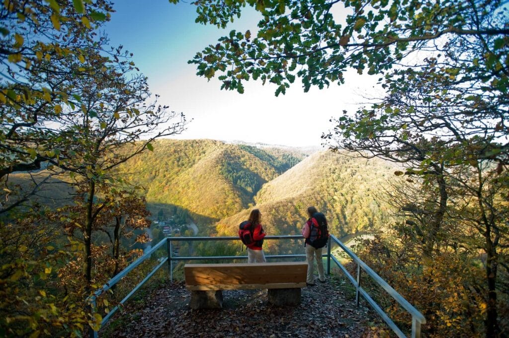 Zwei Frauen in Wanderkleidung mit Rucksäcken stehen auf einem Aussichtspunkt über einem bewaldeten Flusstal. Zwischen dem Betrachter und den Frauen steht eine Holzbank. Einige der Blätter an den Bäumen links und rechts der Wanderinnen zeigen bereits Herbstfarben.