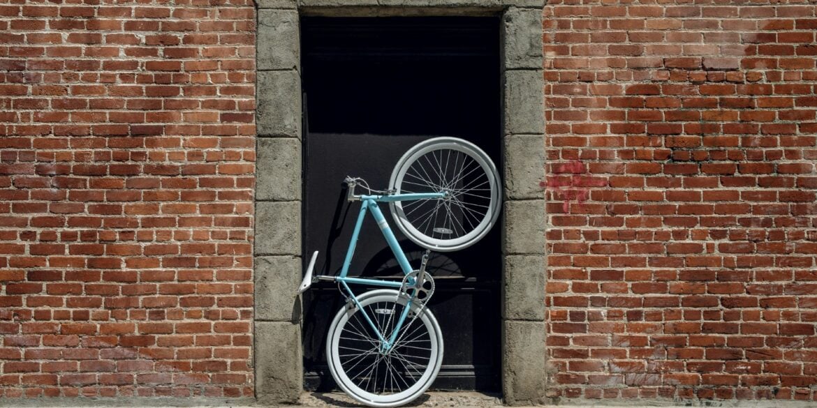 Im Hauseingang eines aus roten Ziegel errichteten Gebäudes steht aufrecht ein sportliches Fahrrad mit einem türkis-blauen Rahmen.