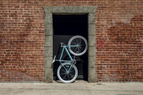 Im Hauseingang eines aus roten Ziegel errichteten Gebäudes steht aufrecht ein sportliches Fahrrad mit einem türkis-blauen Rahmen.