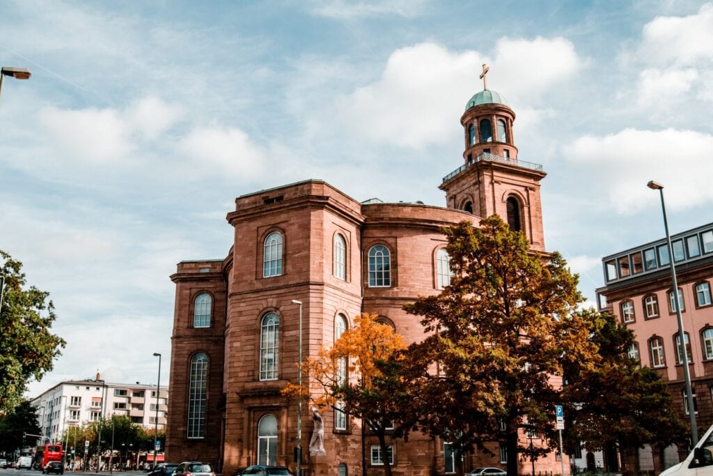 Eine Kirche aus rotem Sandstein; rund um ein rundes Zentralgebäude sind links zwei kleine, quadratische Anbauten angegliedert, rechts ist noch ein Turm mit kupferner Kuppel zu erkennen. Rechts schließt ein weiteres Gebäude an, links der Kirche ist eine Straße. Ein Baum vor der Kirche zeigt, dass es Herbst ist.