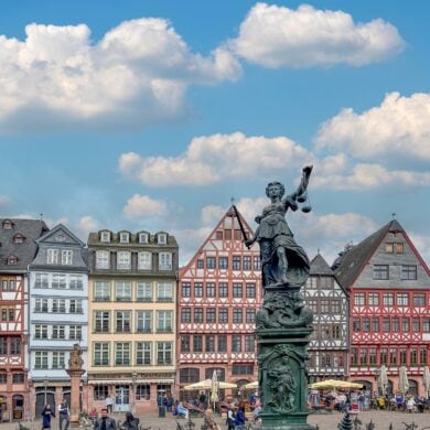Ein Marktplatz mit einer Reihe bunter Fachwerkhäuser. Auf dem Platz sind viele Menschen unterwegs. Im Fokus steht eine hohe Statue der Justitia.