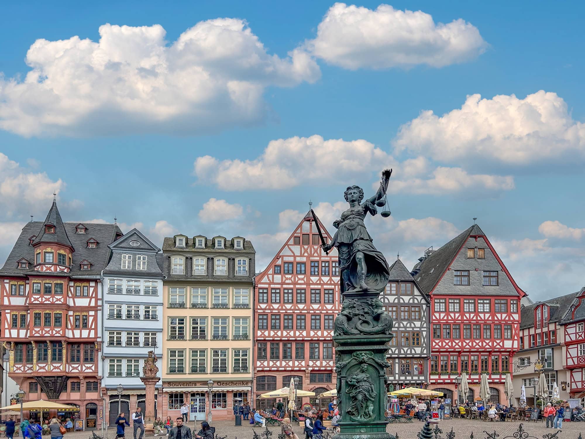 Ein Marktplatz mit einer Reihe bunter Fachwerkhäuser. Auf dem Platz sind viele Menschen unterwegs. Im Fokus steht eine hohe Statue der Justitia.