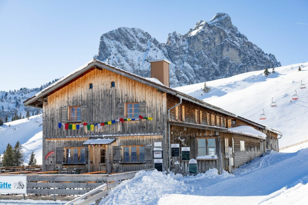 A wooden hut stands in front of a snow-covered Alpine panorama