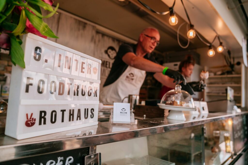 Two people serve food from a food truck. In the foreground is a sign with the inscription "6th Inter / Foodtruck / Rothaus"