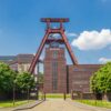 Mittig steht der große Förderturm aus rostrot lackiertem Metall der Zeche Zollverein. Darunter ein geziegeltes Werksgebäude. Vom Betrachter aus führt ein links und rechts von Bäumen gesäumter Weg Richtung Werksgelände