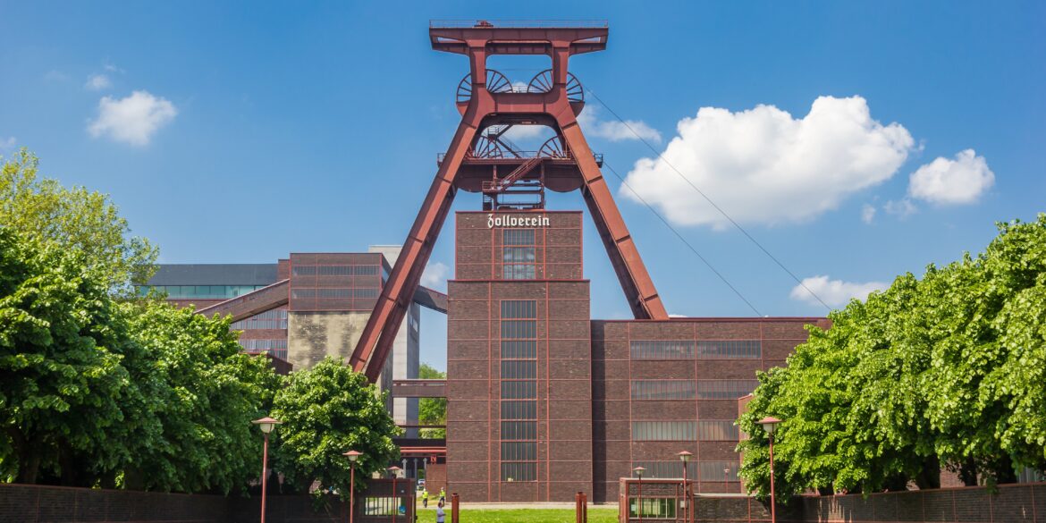 Mittig steht der große Förderturm aus rostrot lackiertem Metall der Zeche Zollverein. Darunter ein geziegeltes Werksgebäude. Vom Betrachter aus führt ein links und rechts von Bäumen gesäumter Weg Richtung Werksgelände