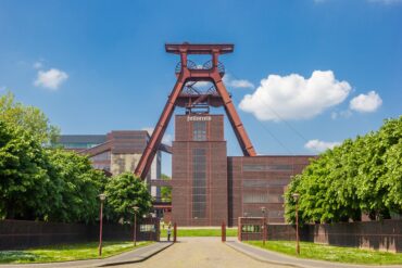 Mittig steht der große Förderturm aus rostrot lackiertem Metall der Zeche Zollverein. Darunter ein geziegeltes Werksgebäude. Vom Betrachter aus führt ein links und rechts von Bäumen gesäumter Weg Richtung Werksgelände