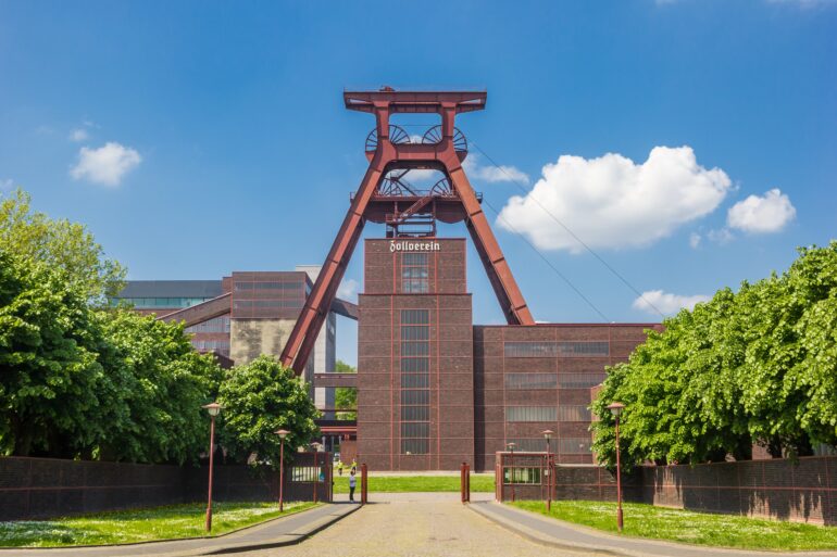 Mittig steht der große Förderturm aus rostrot lackiertem Metall der Zeche Zollverein. Darunter ein geziegeltes Werksgebäude. Vom Betrachter aus führt ein links und rechts von Bäumen gesäumter Weg Richtung Werksgelände