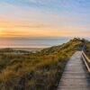 Ein Holzsteg führt vom Betrachter weg durch eine dicht bewachsene, grüne Dünenlandschaft, mit sanft geschwungenen Kuppen. Etwa mittig im Bild wird teilweise das Meer mit Horizont sichtbar, links oben im Bild scheint ein goldener Sonnenuntergang durch eine dünne Wolkendecke und taucht alles in eine sanftes Licht.