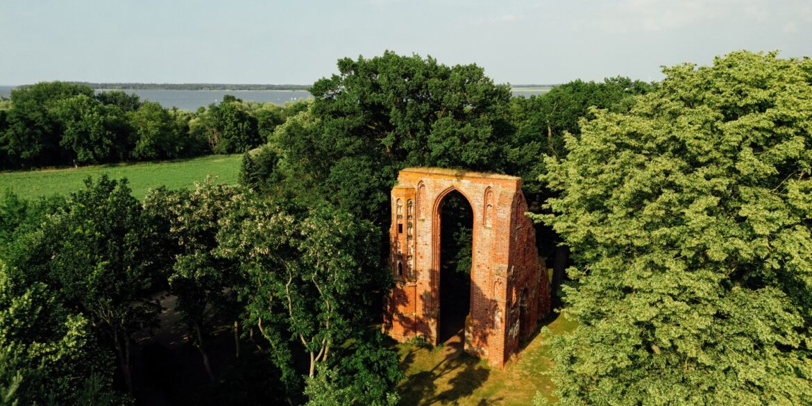 Blick der Drohne auf ein mehrere Meter hohes Portal einer Backsteinkirche, das in einem kleinen Wald steht und von der Sonne angestrahlt wird. Bis auf das Portal ist von der Kirche nichts mehr übrig.