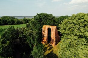 Blick der Drohne auf ein mehrere Meter hohes Portal einer Backsteinkirche, das in einem kleinen Wald steht und von der Sonne angestrahlt wird. Bis auf das Portal ist von der Kirche nichts mehr übrig.