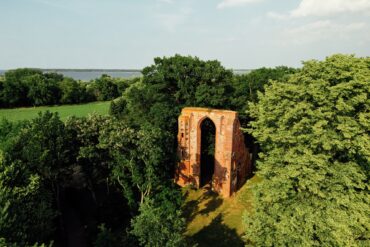Blick der Drohne auf ein mehrere Meter hohes Portal einer Backsteinkirche, das in einem kleinen Wald steht und von der Sonne angestrahlt wird. Bis auf das Portal ist von der Kirche nichts mehr übrig.