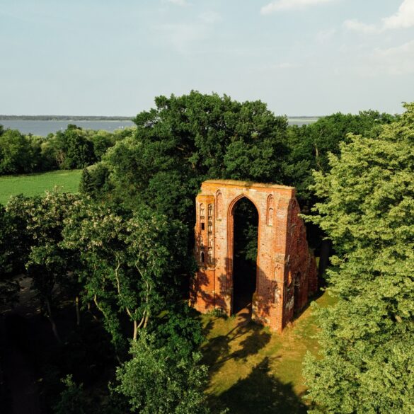 Blick der Drohne auf ein mehrere Meter hohes Portal einer Backsteinkirche, das in einem kleinen Wald steht und von der Sonne angestrahlt wird. Bis auf das Portal ist von der Kirche nichts mehr übrig.