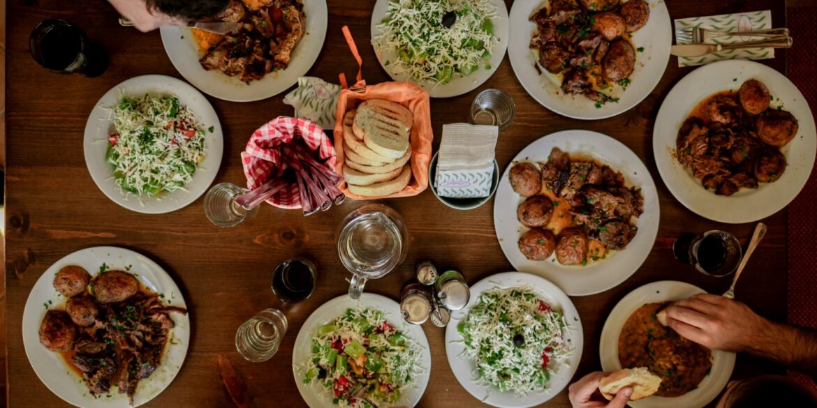 Auf einem Holztisch stehen zahlreiche unterschiedliche Speisen und einige Getränke, der Tisch ist genau von oben fotografiert. Geradeso erkennt man am Rand einige Menschen, die am Tisch sitzen.