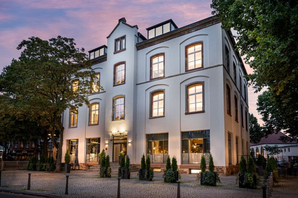 Ein dreistöckiges, weißes Gebäude mit fünf Reihen Fenstern, links und rechts von zwei hohen Laubbäumen flankiert. Das unterste Stockwerk ist durch bodentiefe Fenster von außen einsehbar. Vor dem Gebäude liegt ein gepflasterter Platz. Es ist Abend, das Gebäude ist leicht in gelbes Licht getaucht.