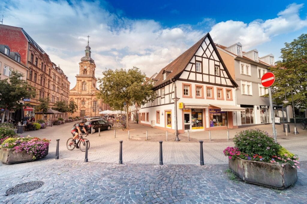 Eine Straßenszene in Saarbrücken; von rechts führt eine gepflasterte Straße in einer Kurve Richtung Bildhintergrund, ein Radler ist auf der Straße nach rechts unterwegs. Rechts im Bild ein Eckhaus, dessen Obergeschoss mit Fachwerk verkleidet ist, rechts davon ein graues, dreigeschossiges Haus. Links im Bild führt die Straße auf eine romanische Sandsteinkirche zu, links davon einige Mehrfamilienhäuser mit rötlich-brauner Fassade. Die dem Betrachter zugewandte Straßenseite ist mit einigen steinernen Blumenkübeln versehen.