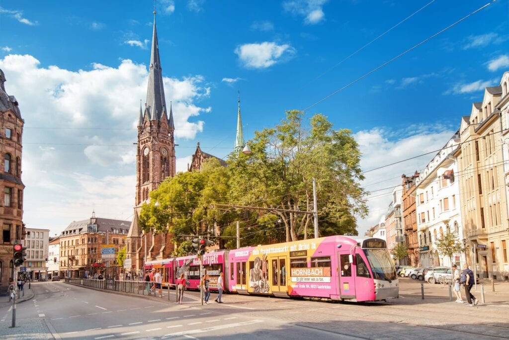An einer zweispurigen Straße fährt eine Straßenbahn mit pinkem Werbeaufdruck nach rechts aus dem Bild heraus. An der Haltestelle auf der gegenüberliegenden Seite stehen einige Menschen. Rechts hinter der Bahn führt eine Straße mit gründerzeitlichen Mehrfamilienhäusern in den Bildhintergrund. Mittig hinter der Straßenbahn einige Laubbäume, hinter denen ein Kirchturm in den Himmel ragt. Alles scheint sehr sommerlich.