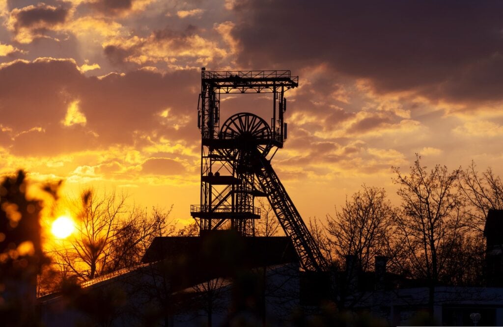 Vor der strahlend untergehenden Sonne zeichnet sich die Silhouette eines Bergwerk-Förderturms mit dem charakteristischen Förderrad ab. Rund um den Turm einige Bäume ohne Blätter, der Himmel ist golden gefärbt.