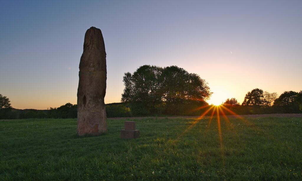 Auf einer weiten Wiese steht ein mehrere Meter hoher Monolith. Im Hintergrund einige Bäume und ein Hügel, hinter dem die Abendsonne untergeht.