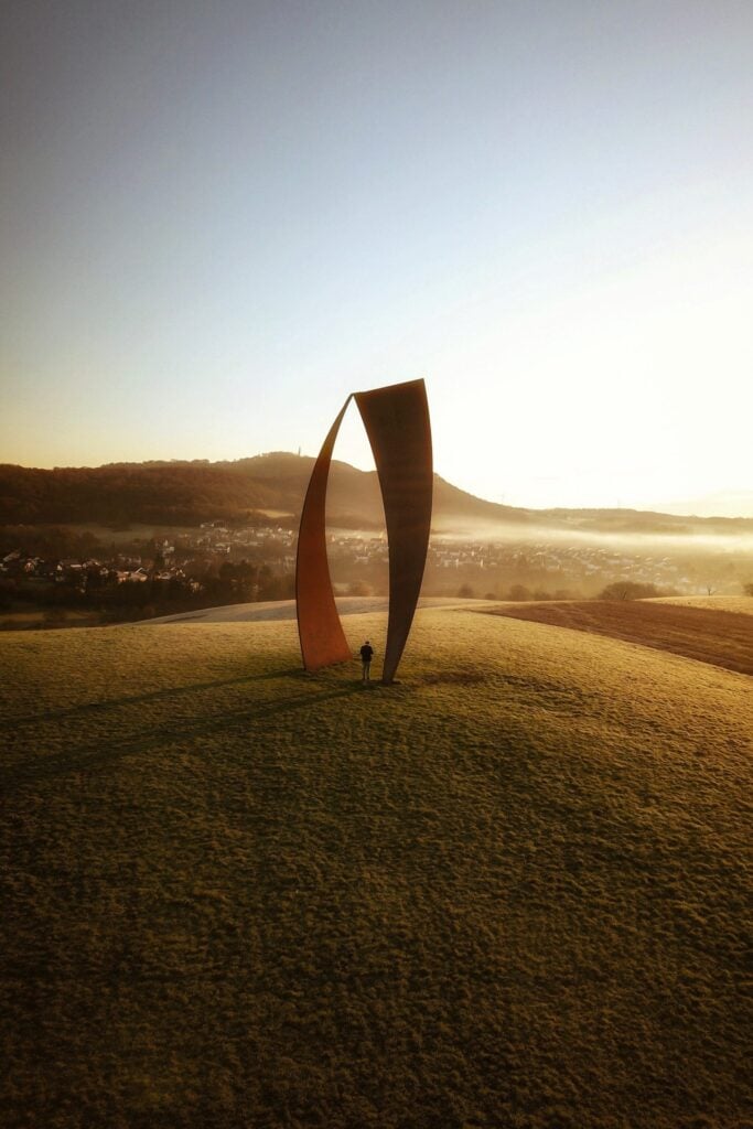 Auf einer Hügelkuppe steht eine mehrere Meter hohe Kunstinstallation aus Metall; links ein Bogen, der sich nach oben verjüngt, rechts ein Bogen, der nach unten verjüngt. Beide treffen sich am oberen Ende. Zwischen den beiden Bögen steht eine Person. Im Hintergrund hinter dem Hügel liegt eine Kleinstadt im Morgennebel, dahinter ein höherer, bewaldeter Hügel.