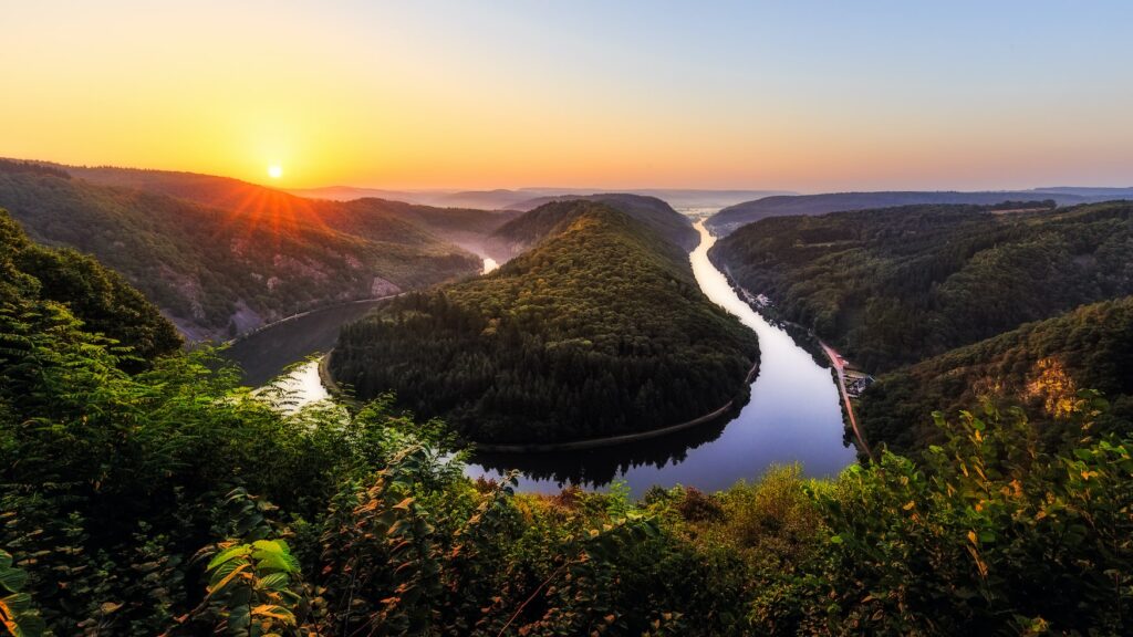 Blick auf eine Flussschleife in U-Form von einem hohen Aussichtspunkt. Die durch die Schleife gebildete Halbinsel ist ebenso bewaldet wie das gesamte Umland. Das Wasser ist spiegelglatt. Links im Hintergrund geht die Sonne rot-orange unter.
