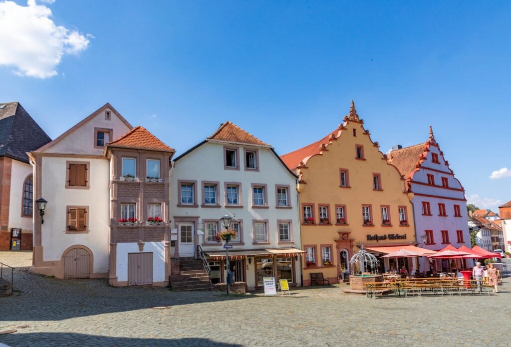 Vier barocke Häuser in hellen Pastellfarben stehen dicht an dicht auf einem nach rechts leicht abschüssigen, gepflasterten Platz. Rechts auf dem Platz stehen einige Bierzeltgarnituren neben einem kleinen Brunnen. Über allem blauer Himmel.