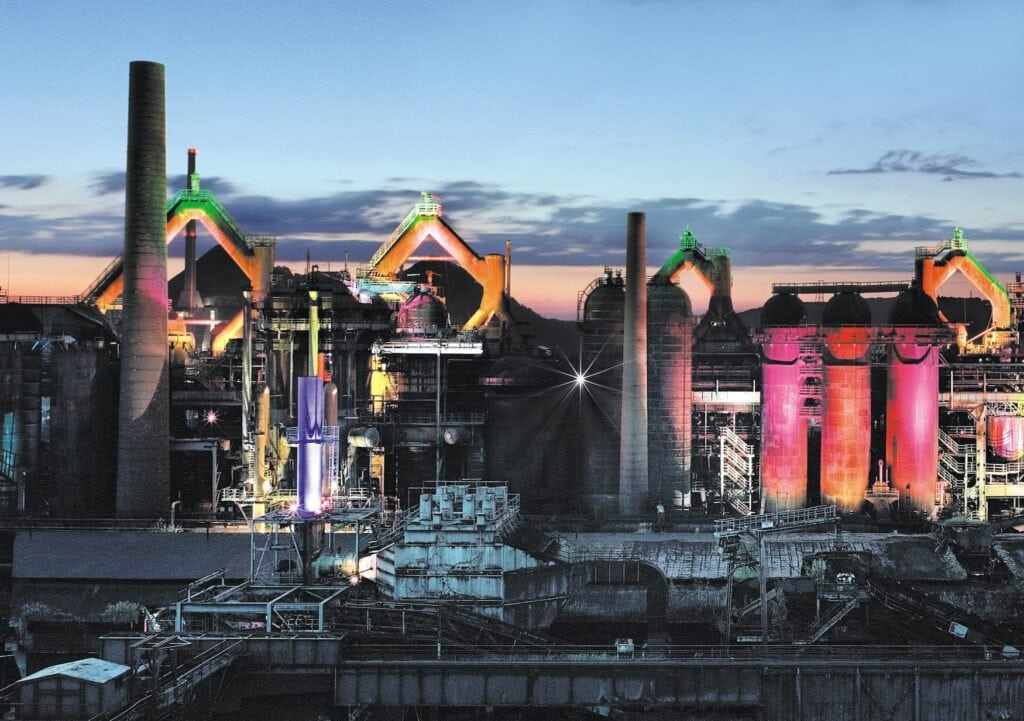 Eine stillgelegte Industrieanlage; links ein hoher Ziegelschronstein, rechts einige Silos, im Vordergrund ein Umspannwerk. Es ist Abend, die Anlage ist von unterschiedlichen Lichtquellen in Farben wie rosa, grün oder gelb angeleuchtet.