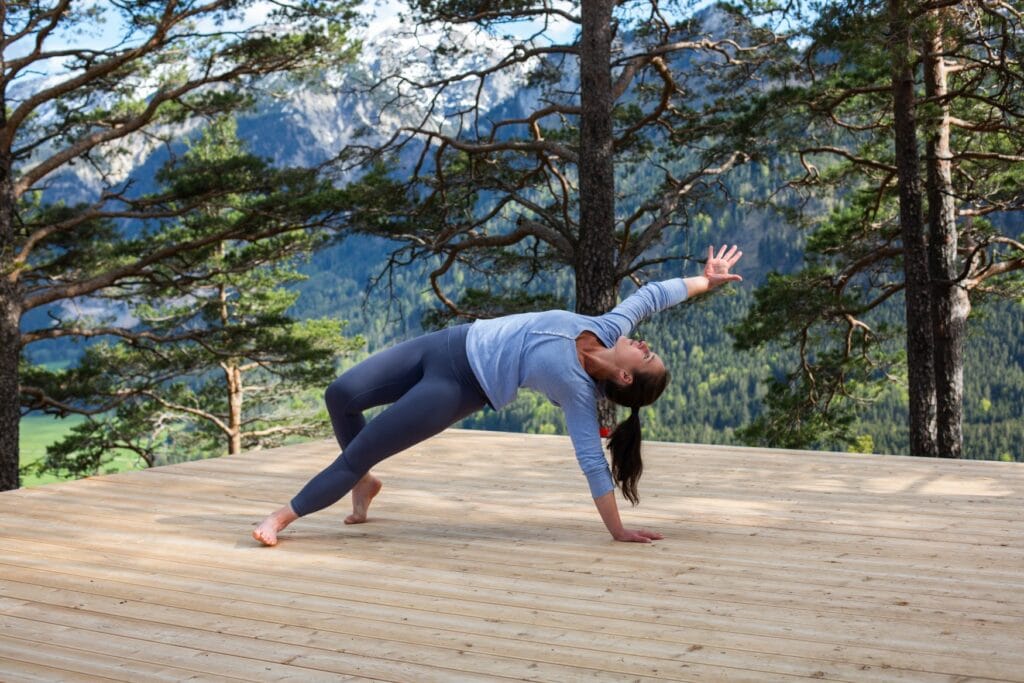 Eine Frau macht Yoga auf einem Holzdeck, im Hintergrund die Alpen