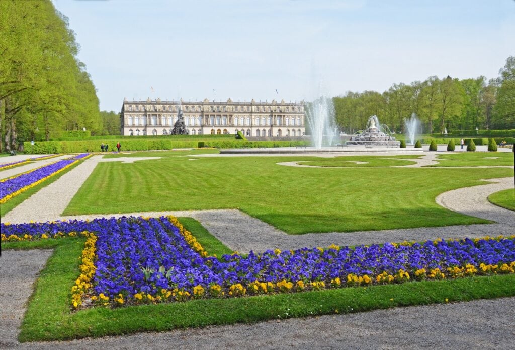 Ein weitläufiger Park mit Blumenbeeten, im Hintergrund das Schloss Herrenchiemsee