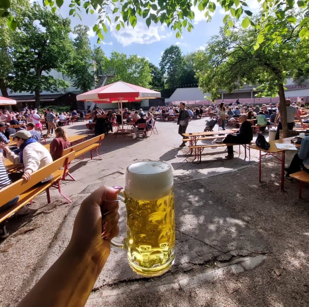 Eine Hand hält vom unteren Bildrand aus einen großen Bierkrug ins Bild. Dahinter ein Platz mit einigen Bäumen und zahlreichen gut belegten Bierzeltgarnituren bei strahlender Sonne