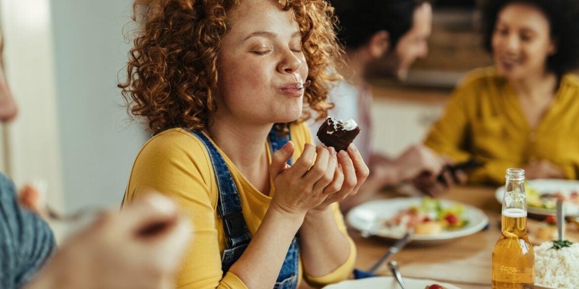 Eine Frau mit roten Locken, einem gelben Shirt und blauer Latzhose beißt von einem Dessert-Gericht ab, während sie an einem gut gefüllten Tisch sitzt