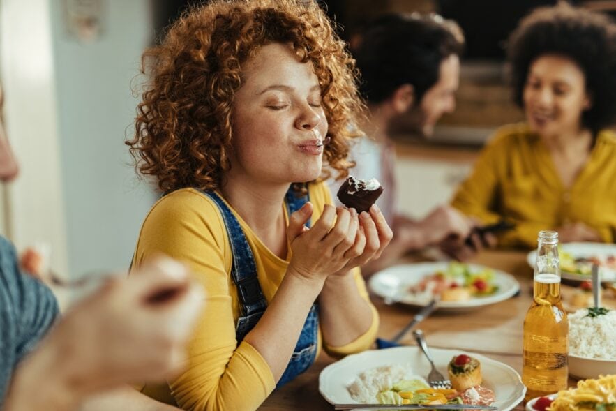 Eine Frau mit roten Locken, einem gelben Shirt und blauer Latzhose beißt von einem Dessert-Gericht ab, während sie an einem gut gefüllten Tisch sitzt