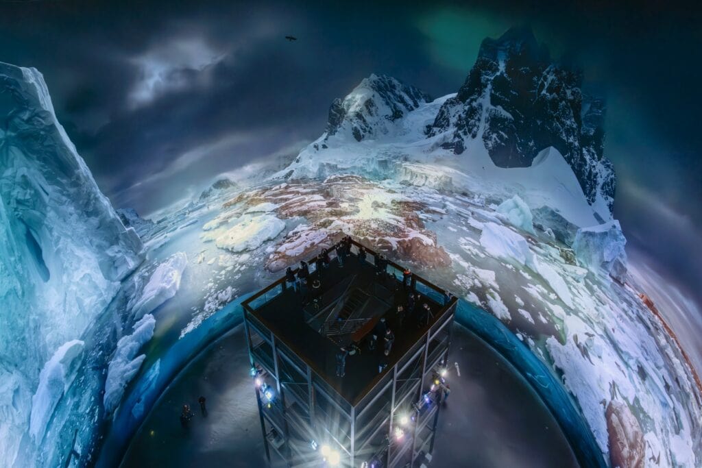 Aufnahme eines Gasometers von innen, ein zylinderförmiger Raum mit einer mittigen Plattform, auf der mehrere Personen stehen. An die Wände wird ein detailreiches Bild der Antarktis projiziert.