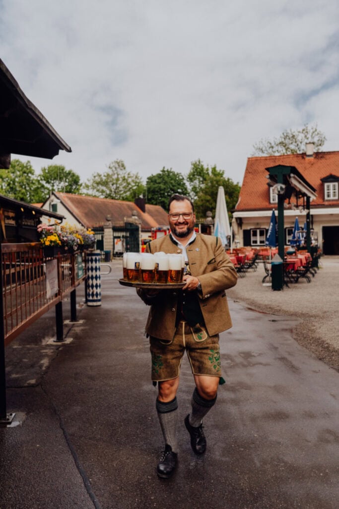 Ein Mann in Tracht trägt mehrere Bierkrüge in einem Biergarten in München