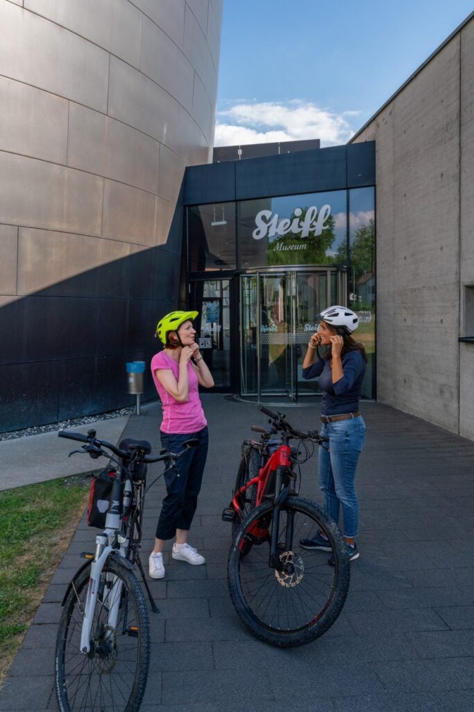 Zwei Radlerinnen setzen sich vor dem Steiff-Museum in Giengen ihre Helme auf.