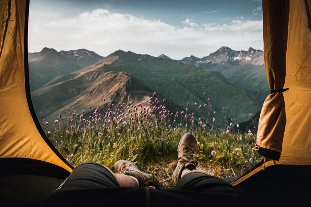 Blick aus einem gelben Zelt auf eine weitläufige Berglandschaft. Unmittelbar vor dem Zelt blühen einige violette Blümchen, zwei Beine in Wanderkleidung der fotografierenden Person sind aus dem Zelt gestreckt.