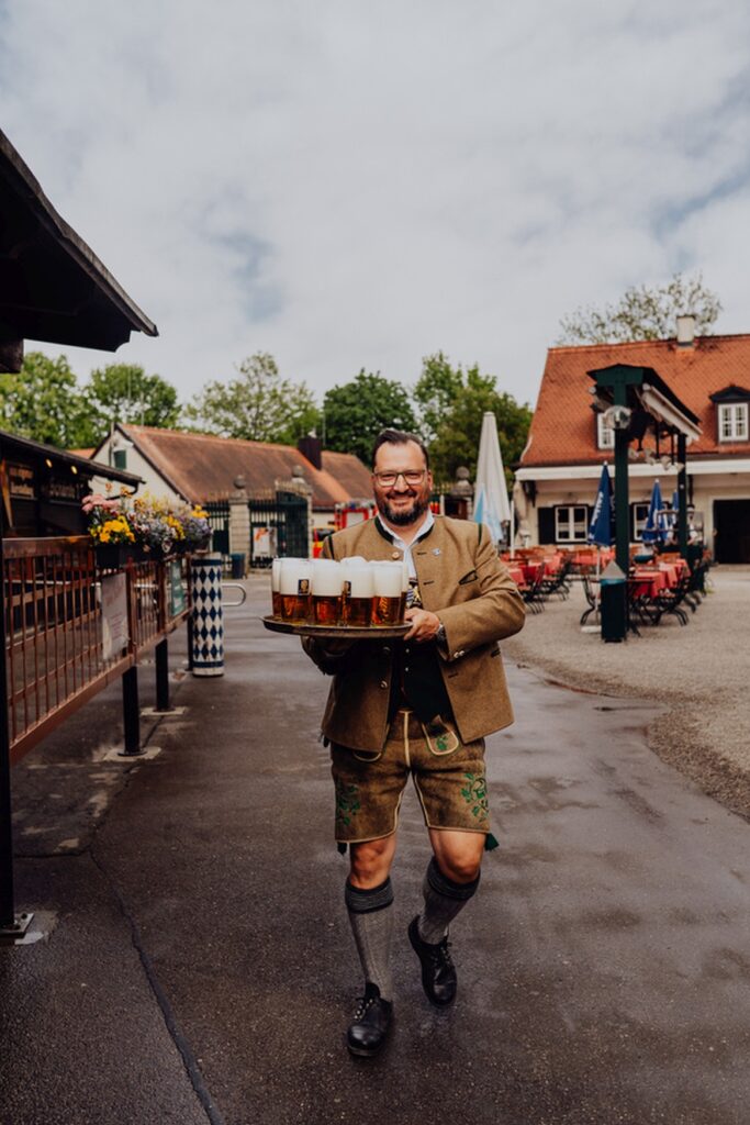 A man in traditional costume carries several beer mugs in a beer garden in Munich