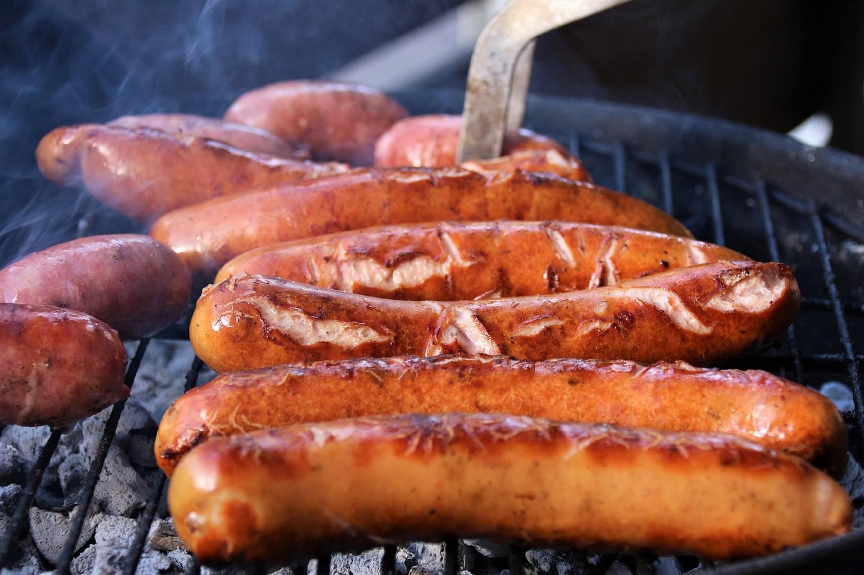 Auf einem rauchenden Grill liegen mehrere rote Bratwürste nebeneinander