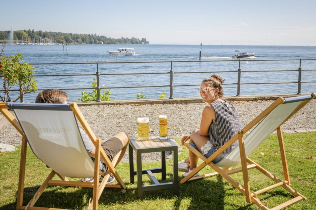 Zwei Personen sitzen in Liegestühlen am Konstanzer Ufer des Bodensees, auf einem Beistelltisch stehen zwei Gläser Bier