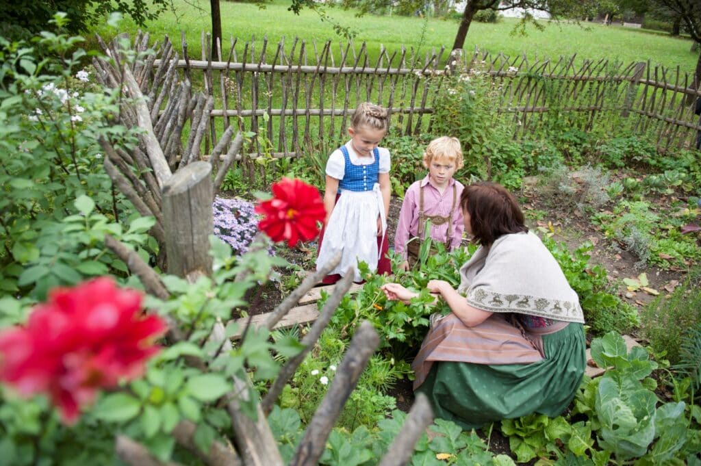 Zwei Kinder lassen sich von einer Frau in historischer Gewandung Kräuter in einem kleinen Garten erklären