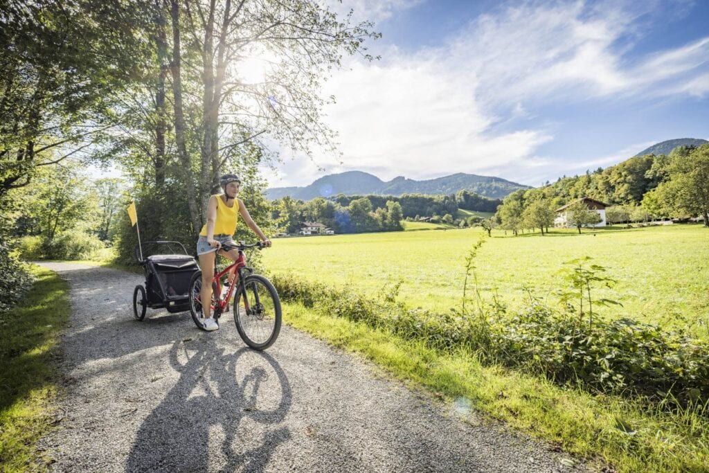 Eine Frau fährt mit Fahrrad und Kinderanhänger entlang eines Fahrradwegs im Chiemsee-Alpenland, daneben eine Wiese, im Hintergrund Berge