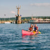 Ein junges Paar paddelt in einem Kanu über den sommerlichen Bodensee vor Konstanz; Im Hintergrund am Ufer ist klar die Statue "Imperia" zu sehen