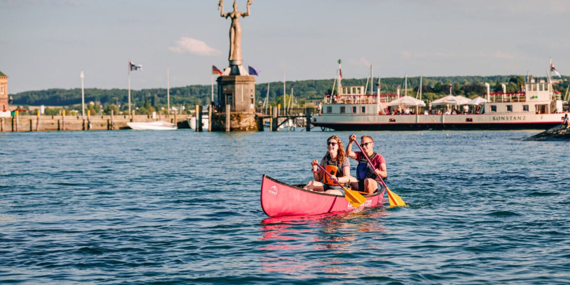 Ein junges Paar paddelt in einem Kanu über den sommerlichen Bodensee vor Konstanz; Im Hintergrund am Ufer ist klar die Statue "Imperia" zu sehen