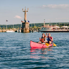 Ein junges Paar paddelt in einem Kanu über den sommerlichen Bodensee vor Konstanz; Im Hintergrund am Ufer ist klar die Statue "Imperia" zu sehen