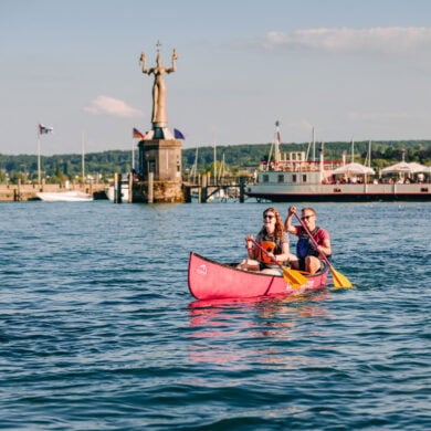 Ein junges Paar paddelt in einem Kanu über den sommerlichen Bodensee vor Konstanz; Im Hintergrund am Ufer ist klar die Statue "Imperia" zu sehen