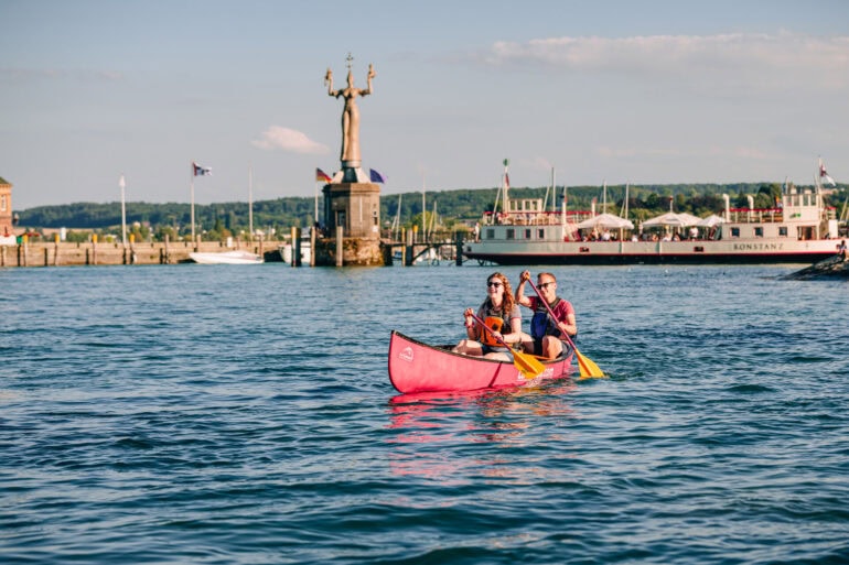 Ein junges Paar paddelt in einem Kanu über den sommerlichen Bodensee vor Konstanz; Im Hintergrund am Ufer ist klar die Statue "Imperia" zu sehen