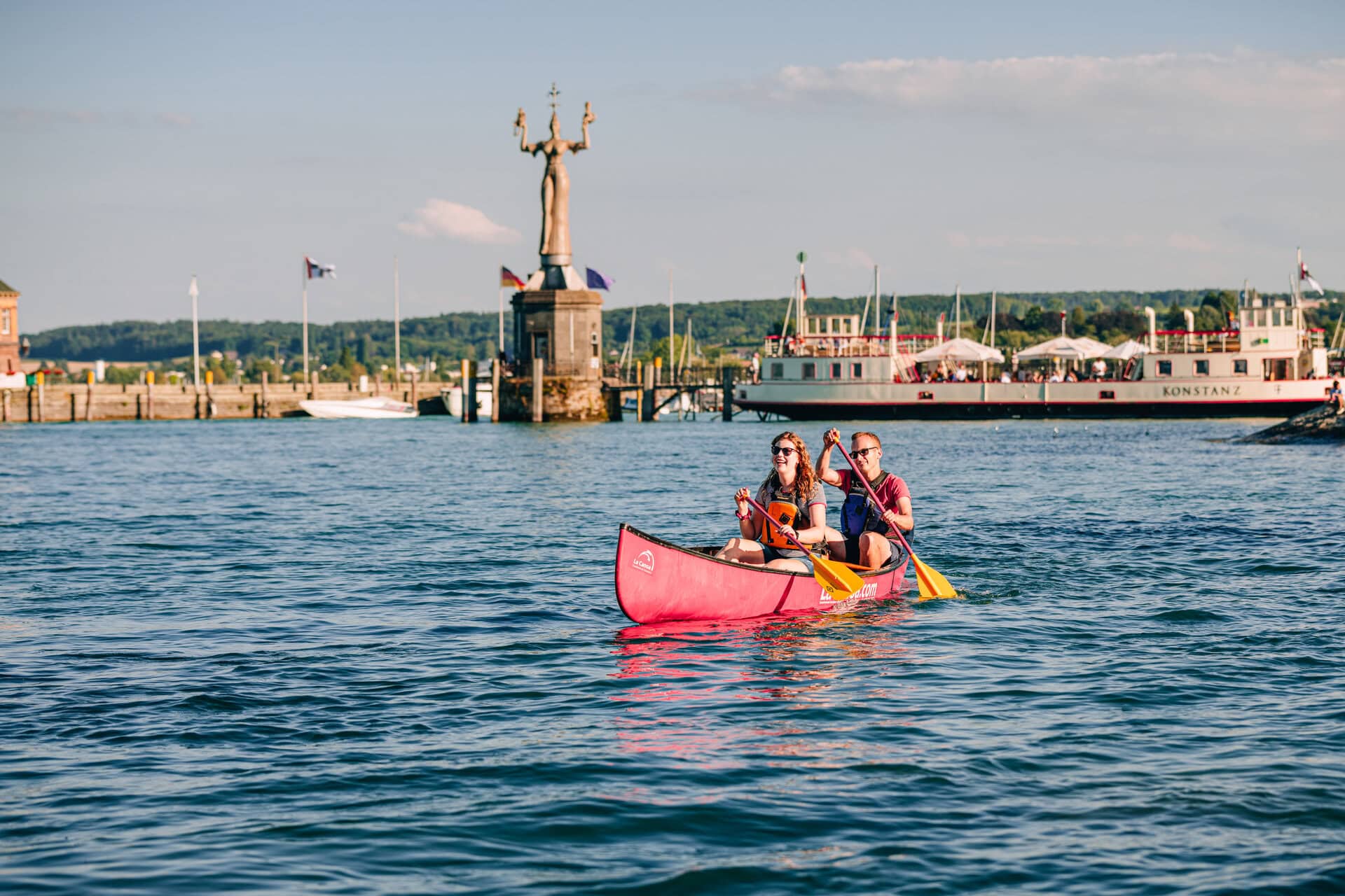 Ein junges Paar paddelt in einem Kanu über den sommerlichen Bodensee vor Konstanz; Im Hintergrund am Ufer ist klar die Statue "Imperia" zu sehen