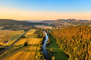 Die Saale aus der Vogelperspektive im Licht der Abendsonne. Links des Flusses liegen Felder, rechts ein Wald. Am Horizont liegt die Leuchtenburg.